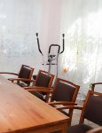 Wnętrze dziennego domu. Duży drewniany stół z tapicerowanymi krzesłami. W tle steper treningowy i dwa duże okna z ażurowymi firanami